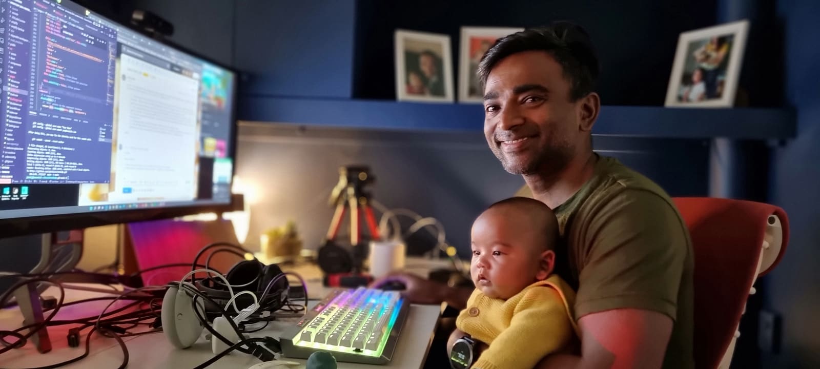 Dr Jubal at his desk with code on the screen and a baby in a yellow cardigan on his lap, eyes on the keyboard