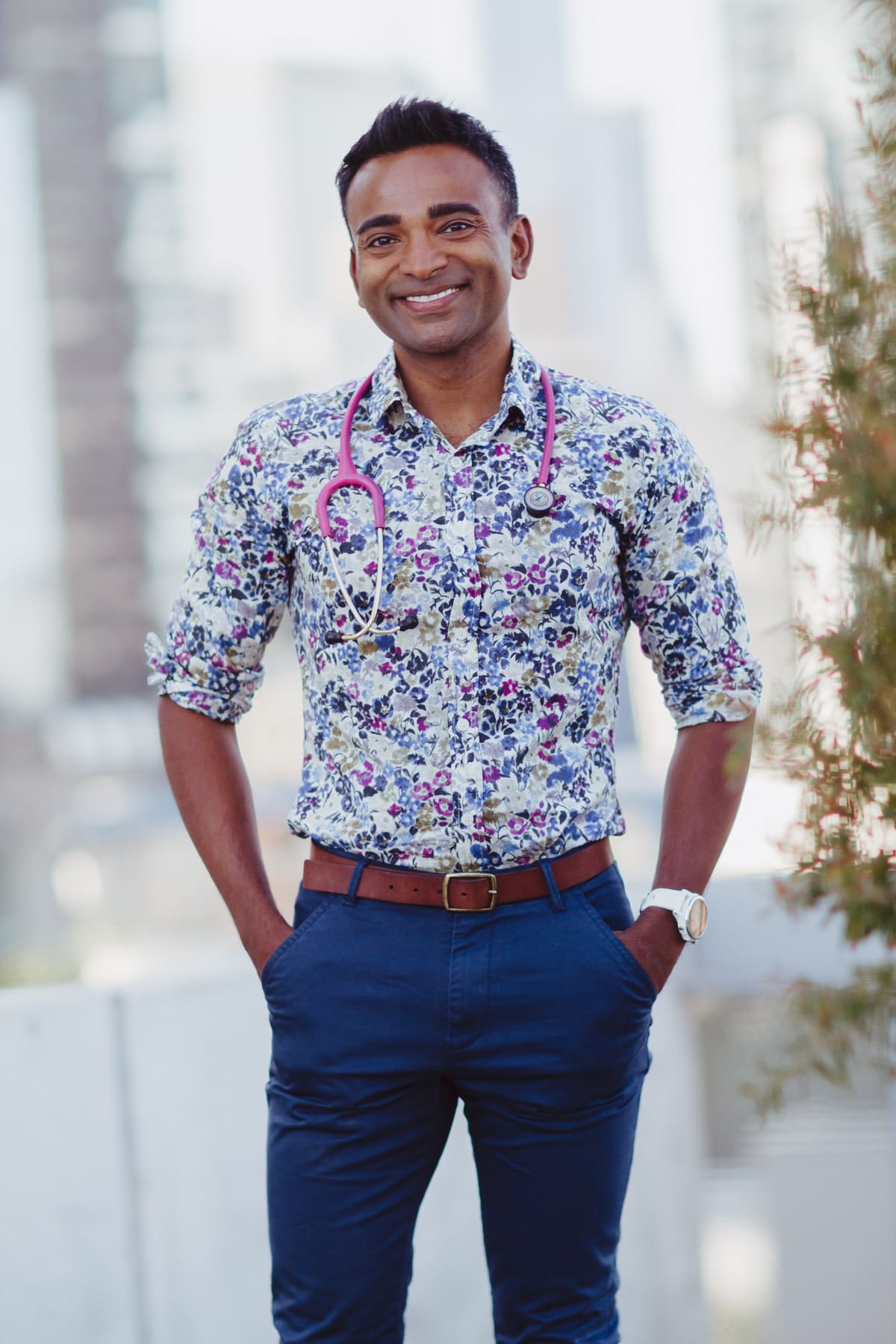 Dr Jubal John standing outdoors in a floral shirt, pink stethoscope around his neck, Melbourne city soft-focus behind