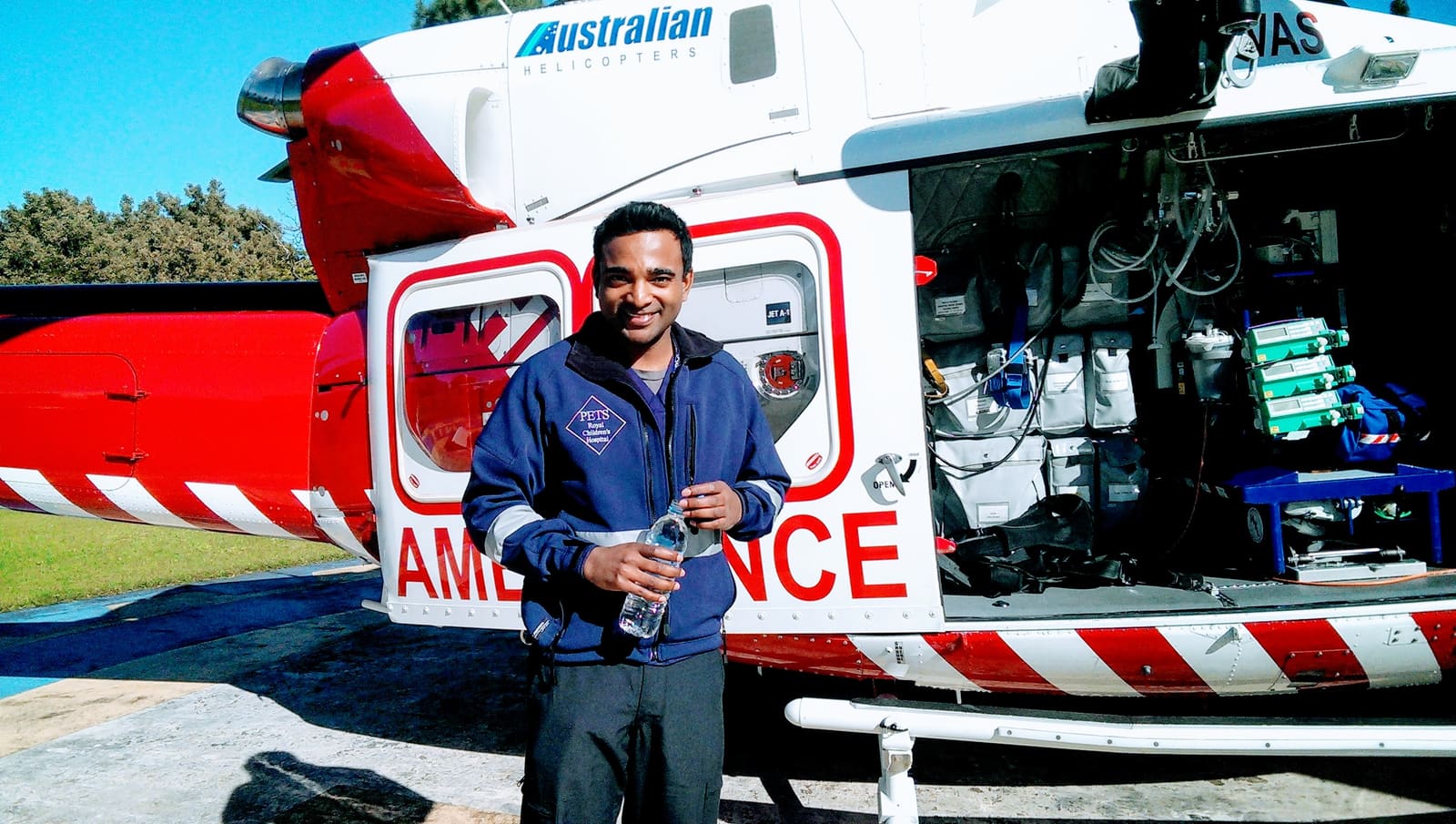A younger Dr Jubal in a Royal Children's Hospital PETS retrieval jacket in front of an air-ambulance helicopter, its medical equipment bay open